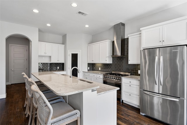 kitchen with arched walkways, stainless steel appliances, a sink, light stone countertops, and wall chimney exhaust hood