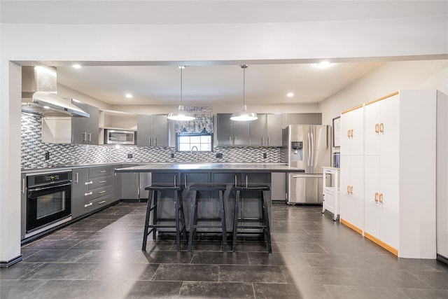 kitchen with wall chimney exhaust hood, appliances with stainless steel finishes, a breakfast bar area, a center island, and gray cabinetry