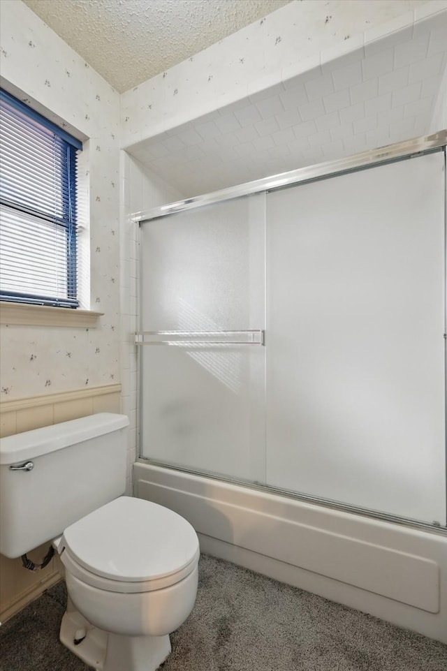 full bathroom featuring wallpapered walls, toilet, shower / bath combination with glass door, a wainscoted wall, and a textured ceiling