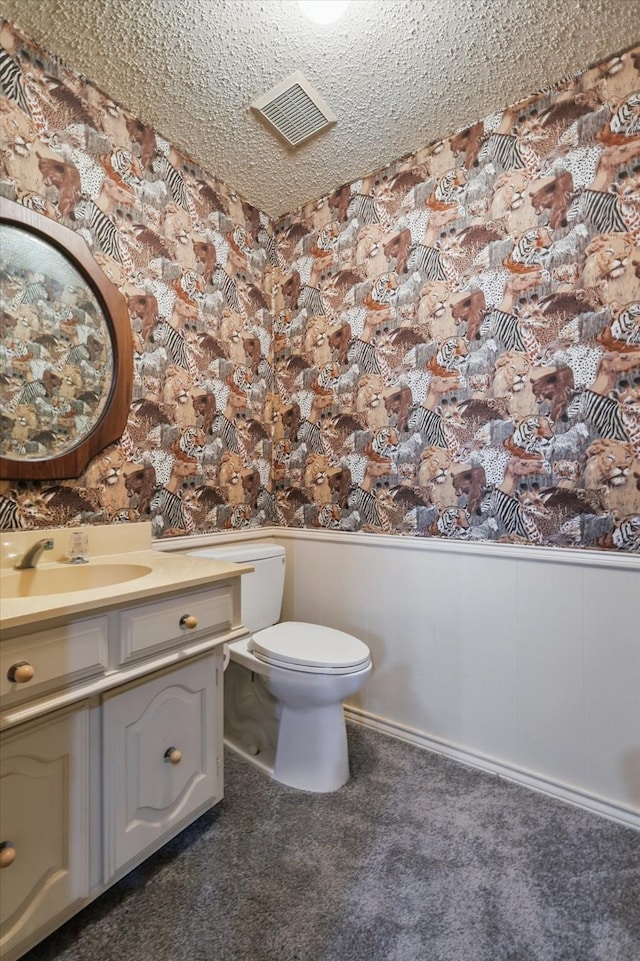 half bathroom featuring wallpapered walls, visible vents, wainscoting, toilet, and a textured ceiling