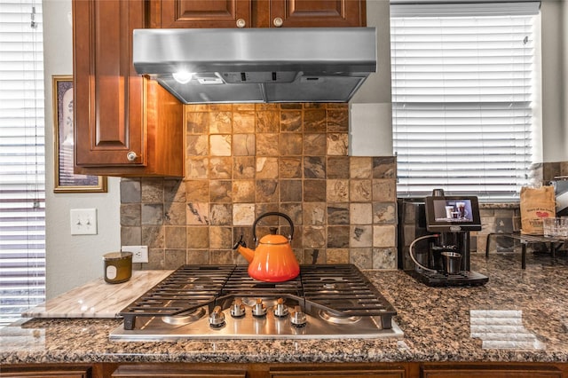 kitchen with dark stone counters, brown cabinets, ventilation hood, stainless steel gas cooktop, and backsplash