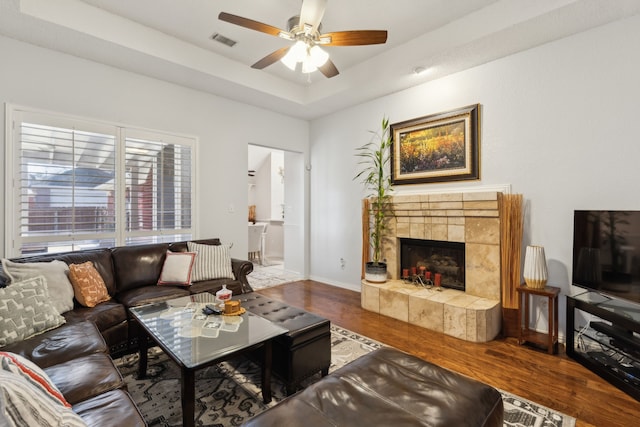 living area featuring a tile fireplace, wood finished floors, visible vents, a ceiling fan, and a raised ceiling