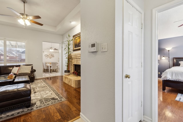 living area featuring a fireplace with raised hearth, a tray ceiling, wood finished floors, and a ceiling fan