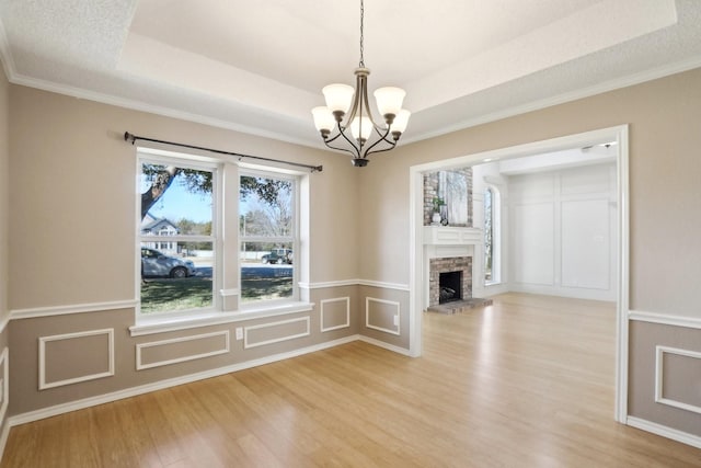 unfurnished dining area featuring a stone fireplace, a decorative wall, light wood finished floors, a raised ceiling, and an inviting chandelier