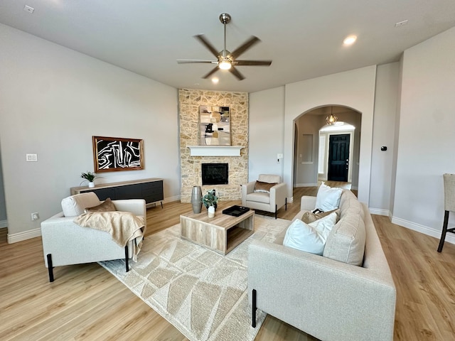 living area featuring light wood-type flooring, arched walkways, a stone fireplace, and a ceiling fan