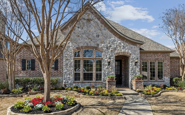 french country inspired facade with roof with shingles, stone siding, and brick siding