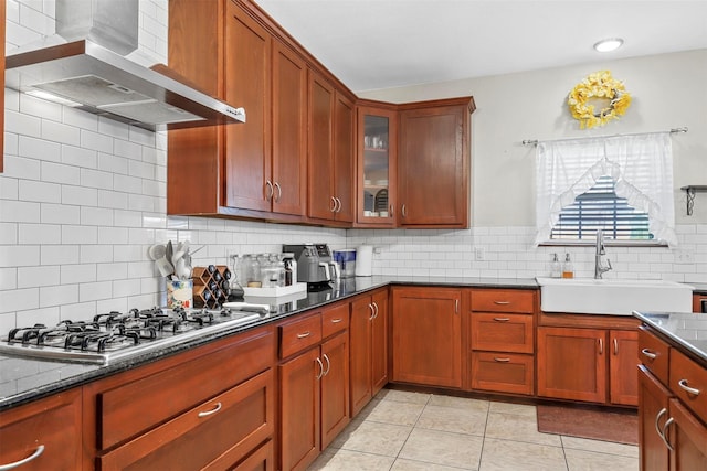 kitchen with stainless steel gas cooktop, a sink, wall chimney range hood, decorative backsplash, and glass insert cabinets