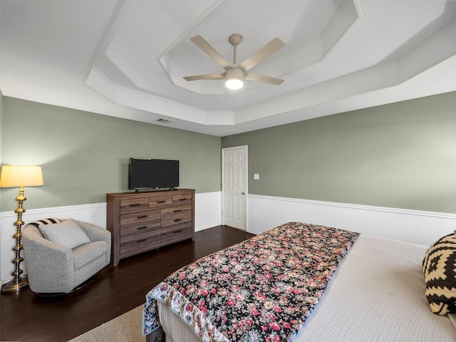 bedroom featuring a tray ceiling, a wainscoted wall, visible vents, a ceiling fan, and wood finished floors