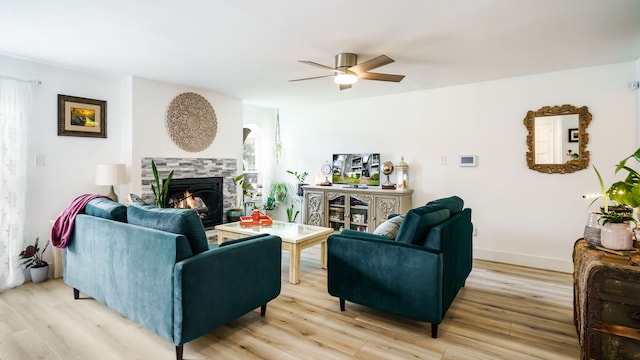 living area featuring light wood-style floors, ceiling fan, a stone fireplace, and baseboards