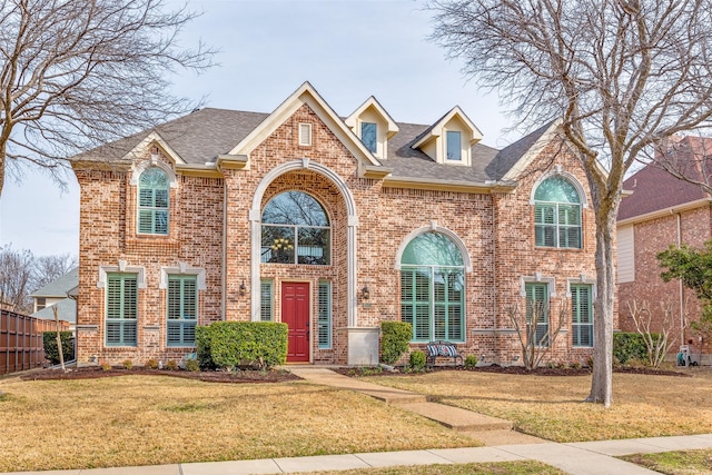 traditional-style home with brick siding, fence, a front lawn, and roof with shingles