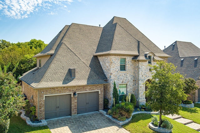 french country style house with a garage, a shingled roof, stone siding, decorative driveway, and a front lawn