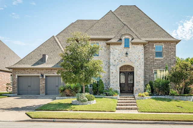 french country inspired facade featuring a shingled roof, stone siding, an attached garage, decorative driveway, and a front yard