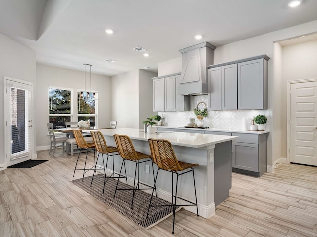 kitchen featuring a kitchen island with sink, gray cabinetry, light countertops, tasteful backsplash, and custom range hood