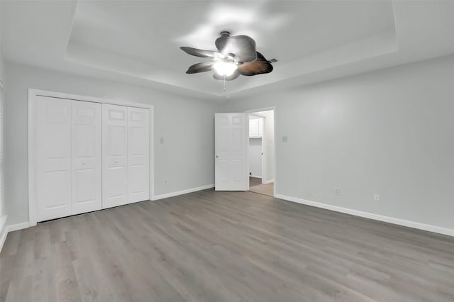 unfurnished bedroom featuring ceiling fan, wood finished floors, baseboards, a closet, and a tray ceiling