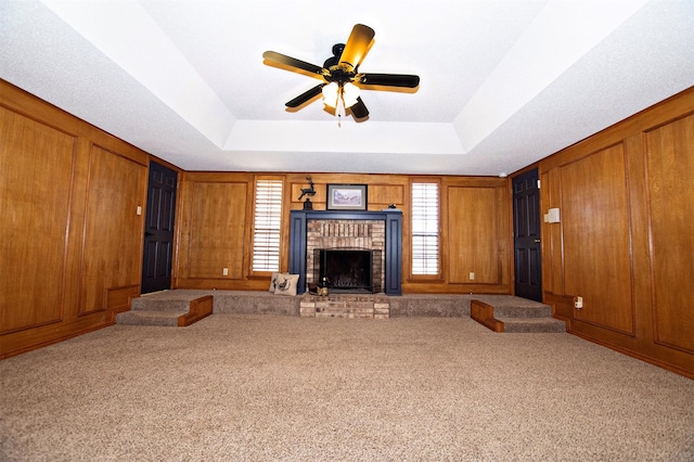 unfurnished living room featuring a brick fireplace, a raised ceiling, carpet flooring, and wooden walls