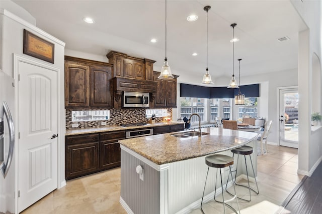 kitchen featuring tasteful backsplash, stainless steel microwave, a sink, an island with sink, and light stone countertops