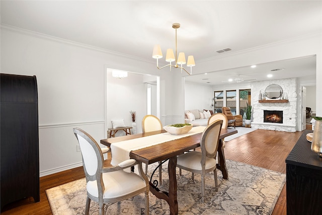 dining area with a notable chandelier, visible vents, ornamental molding, a stone fireplace, and wood finished floors