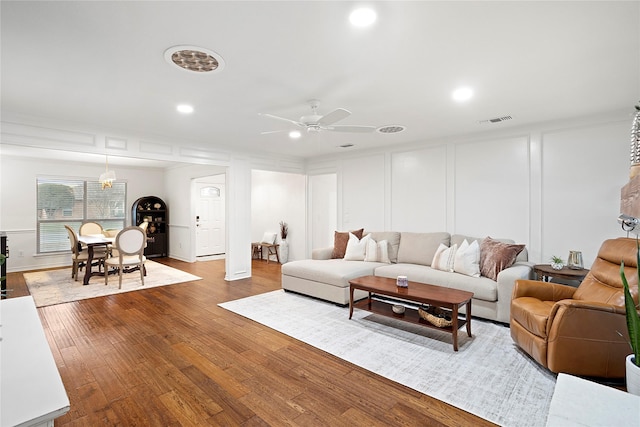 living room featuring a ceiling fan, visible vents, a decorative wall, and wood finished floors