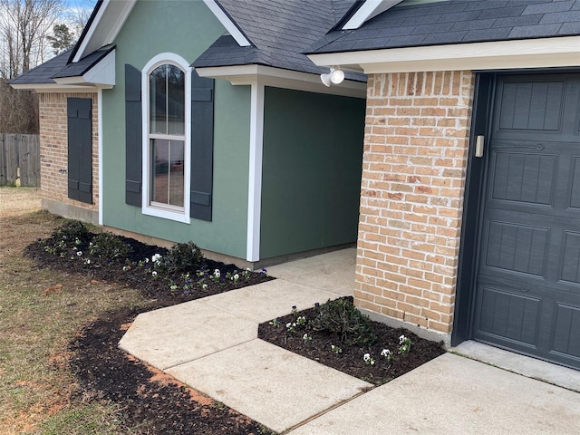 property entrance featuring a garage, brick siding, fence, and roof with shingles