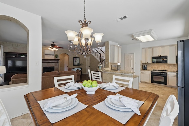 dining room featuring arched walkways, light tile patterned floors, a tile fireplace, ceiling fan with notable chandelier, and visible vents