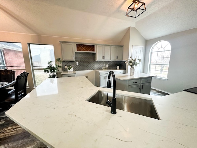 kitchen with gray cabinetry, a sink, vaulted ceiling, light stone countertops, and tasteful backsplash