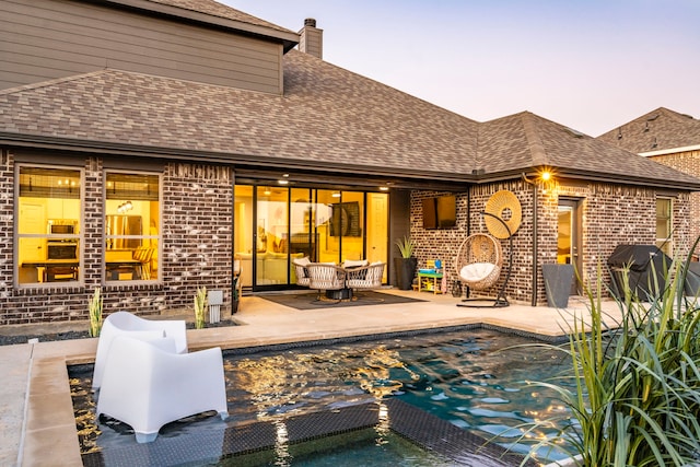 back of property with a shingled roof, brick siding, a patio, and a chimney