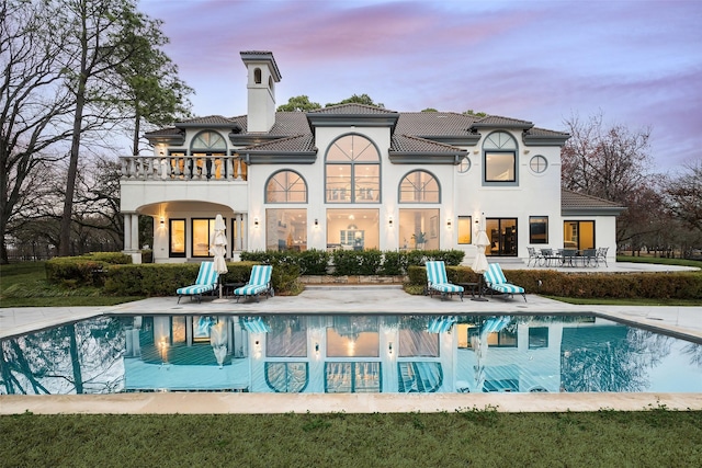 back of property at dusk featuring stucco siding, a chimney, a balcony, a patio area, and an outdoor pool