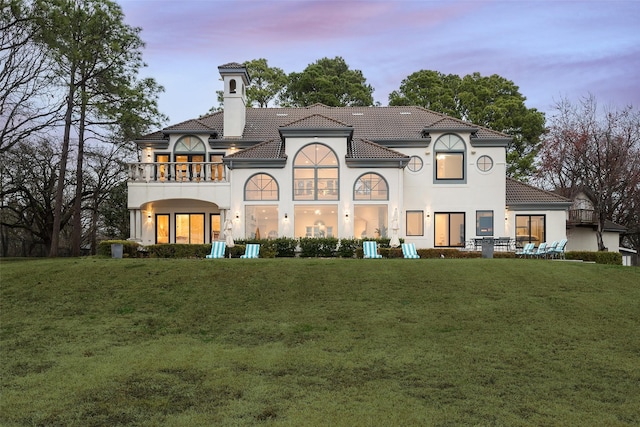 back of house at dusk featuring a balcony, a lawn, a chimney, and a tiled roof