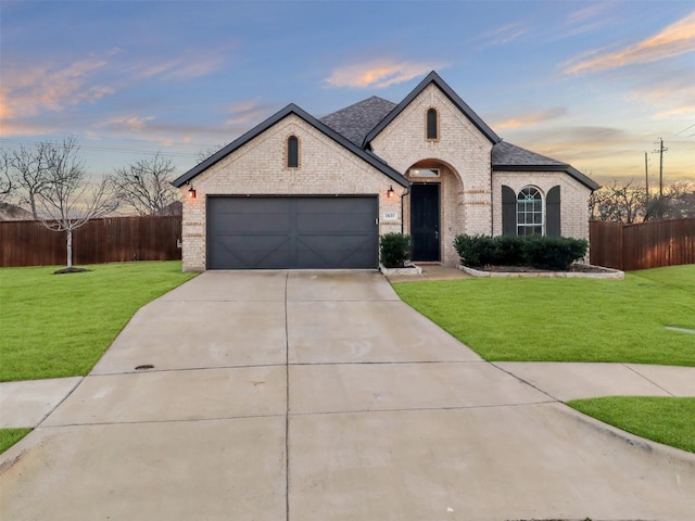 french country inspired facade with concrete driveway, brick siding, a lawn, and fence