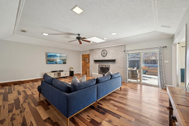 living area featuring a skylight, wood finished floors, visible vents, baseboards, and a brick fireplace