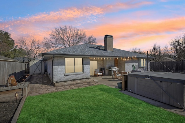 back of property at dusk featuring a fenced backyard, brick siding, a yard, a patio area, and a hot tub