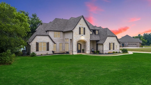 french country style house with a front yard, stone siding, central AC, and roof with shingles