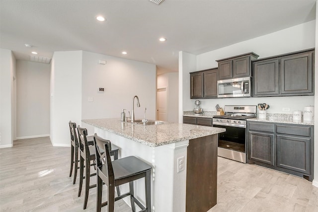 kitchen with a breakfast bar, appliances with stainless steel finishes, a sink, an island with sink, and light stone countertops