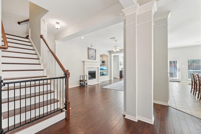 entryway with dark wood finished floors, decorative columns, a fireplace with flush hearth, ceiling fan, and stairs