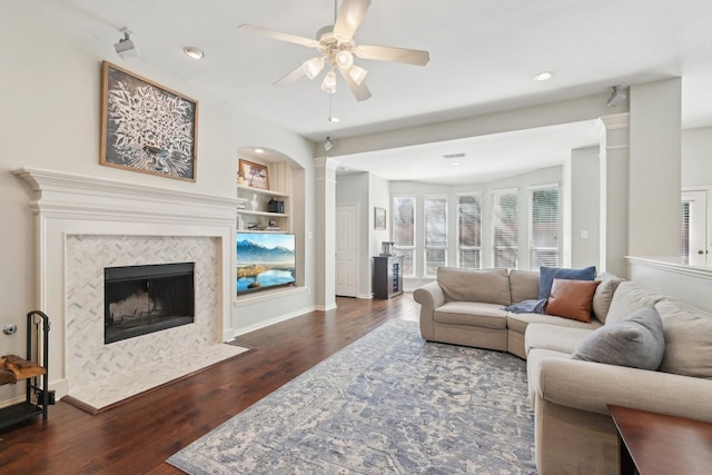 living area featuring dark wood-style flooring, a ceiling fan, built in features, a tiled fireplace, and ornate columns