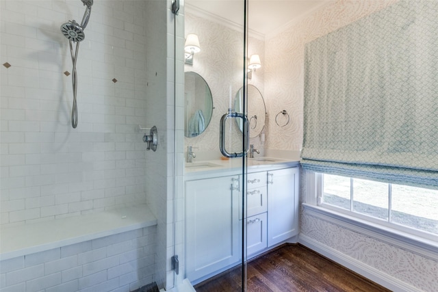 bathroom with wood finished floors, a sink, tiled shower, double vanity, and wallpapered walls