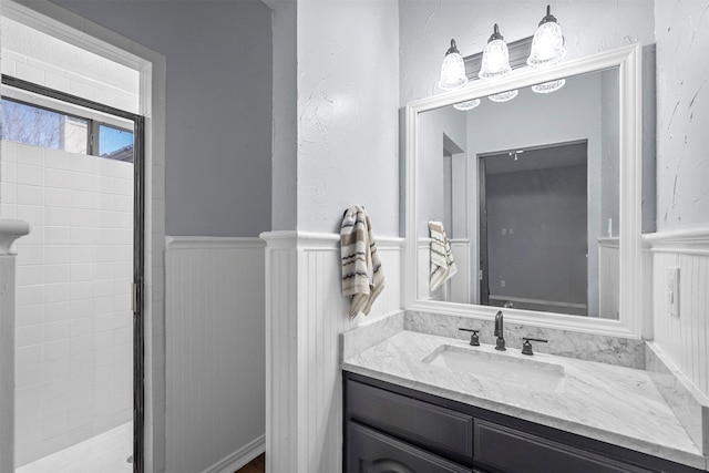 full bathroom featuring a wainscoted wall, a textured wall, vanity, and tiled shower