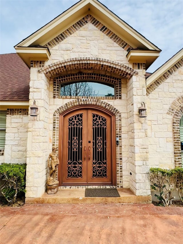 view of exterior entry with stone siding, french doors, and roof with shingles