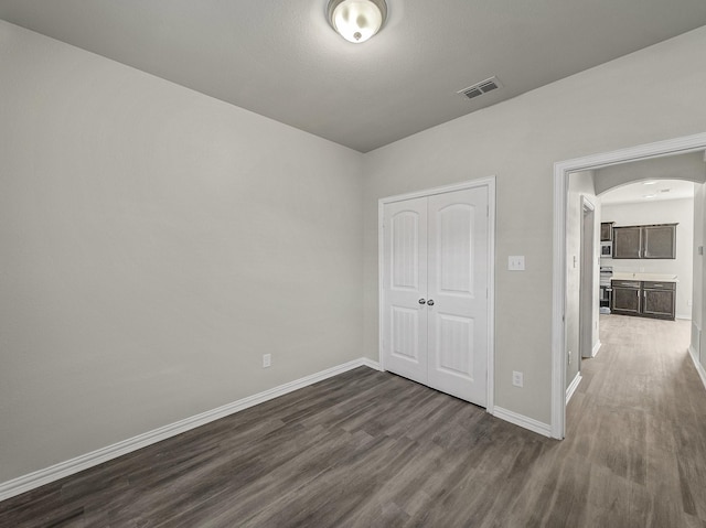 unfurnished bedroom featuring arched walkways, dark wood-style flooring, a closet, visible vents, and baseboards