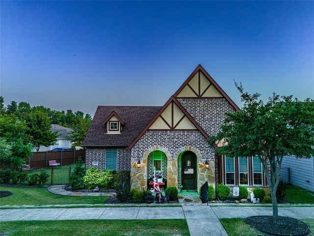 english style home featuring roof with shingles, stucco siding, a front yard, fence, and stone siding