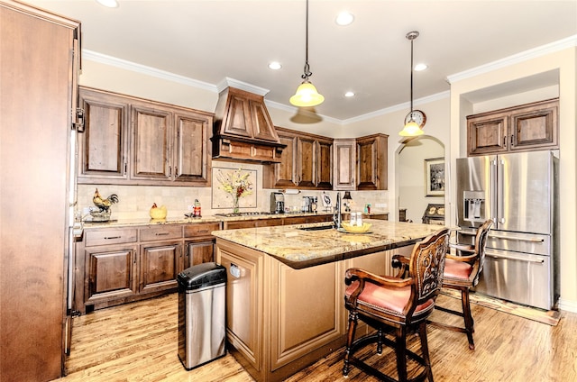 kitchen featuring a center island with sink, stainless steel appliances, a sink, light stone countertops, and premium range hood