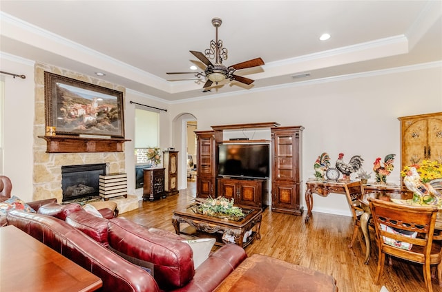 living area featuring a raised ceiling, a ceiling fan, ornamental molding, wood finished floors, and a stone fireplace