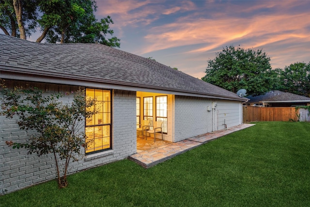 back of house at dusk featuring a yard, a shingled roof, fence, and brick siding