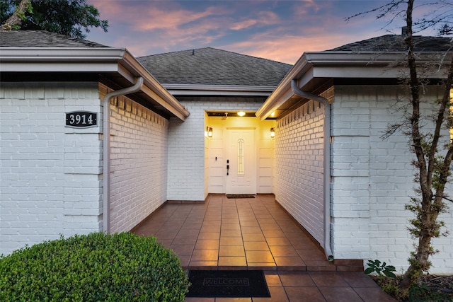 exterior entry at dusk featuring brick siding, a patio, and roof with shingles