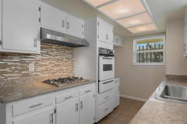 kitchen with white oven, stainless steel gas cooktop, backsplash, white cabinets, and under cabinet range hood