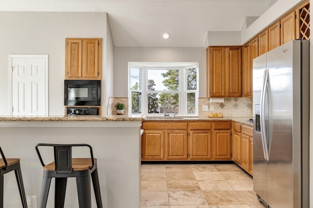 kitchen with stainless steel fridge with ice dispenser, backsplash, light stone countertops, black microwave, and a sink