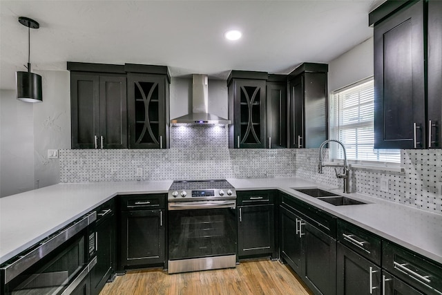 kitchen featuring light countertops, appliances with stainless steel finishes, a sink, wall chimney range hood, and dark cabinetry