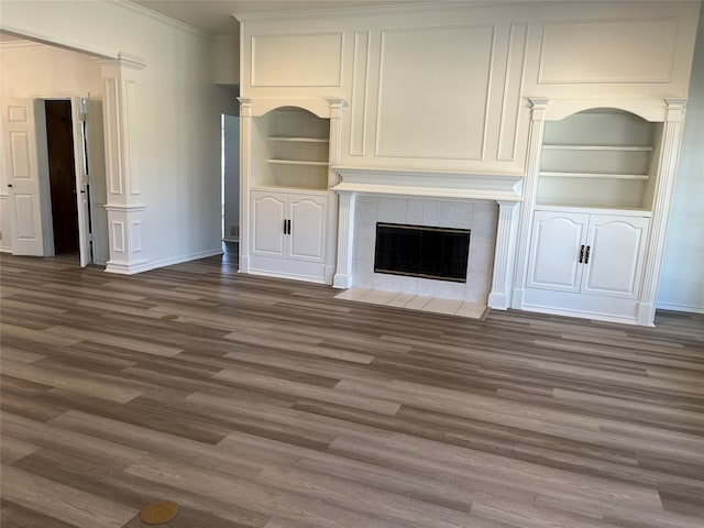 unfurnished living room with a tile fireplace, wood finished floors, built in features, ornamental molding, and ornate columns