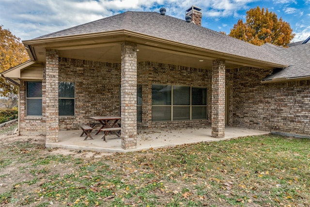 back of house with a chimney, brick siding, roof with shingles, and a patio area