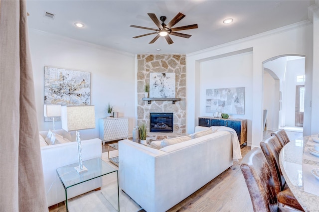 living room featuring arched walkways, crown molding, light wood finished floors, visible vents, and a stone fireplace